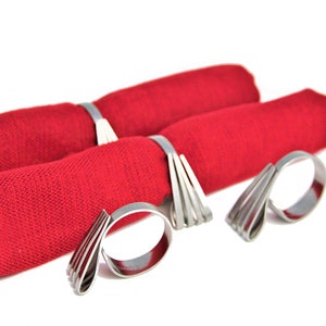 May include: Three silver napkin rings with a unique, modern design. The rings are made of thin, flat metal strips that are bent and layered to create a decorative, geometric pattern. The rings are shown holding red napkins.