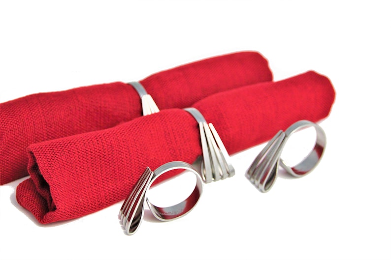 May include: Three silver napkin rings with a unique, modern design. The rings are made of thin, flat metal strips that are bent and layered to create a decorative, geometric pattern. The rings are shown holding red napkins.