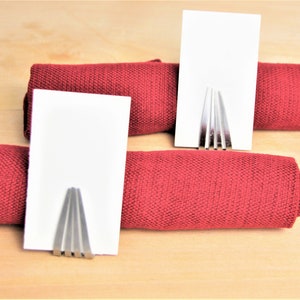 May include: Two silver metal place card holders with blank white cards on top of red fabric napkin rolls.