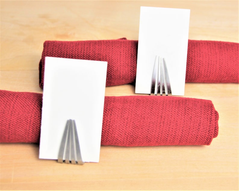 May include: Two silver metal place card holders with blank white cards on top of red fabric napkin rolls.