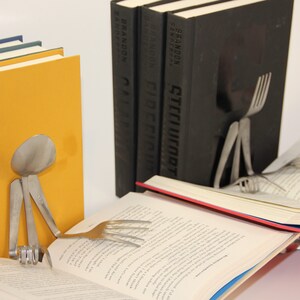 Puede incluir: Tres libros de tapa dura negros con texto blanco en los lomos. Los libros se titulan "Stellucort", "Cideficut" y "Nav", todos de Brandon Sandeson. Un tenedor y una cuchara de plata se utilizan como sujetalibros.
