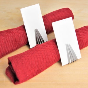 May include: Two red fabric napkins are rolled up and held together with a silver fork napkin ring. Each napkin ring has a blank white card placed in it.