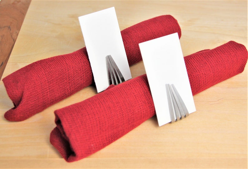 May include: Two red fabric napkins are rolled up and held together with a silver fork napkin ring. Each napkin ring has a blank white card placed in it.