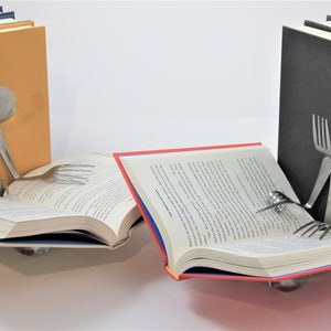 Puede incluir: Dos sujetalibros hechos de cubiertos est&aacute;n sosteniendo dos libros abiertos. Los sujetalibros tienen forma de personas leyendo. Un sujetalibros sostiene un libro con una cubierta amarilla y el otro sujetalibros sostiene un libro con una cubierta roja.