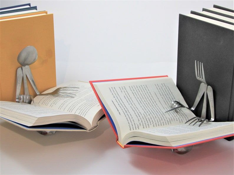 Puede incluir: Dos sujetalibros hechos de cubiertos est&aacute;n sosteniendo dos libros abiertos. Los sujetalibros tienen forma de personas leyendo. Un sujetalibros sostiene un libro con una cubierta amarilla y el otro sujetalibros sostiene un libro con una cubierta roja.