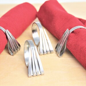 May include: Four silver napkin rings made of stacked metal strips. The rings are shown on red napkins.