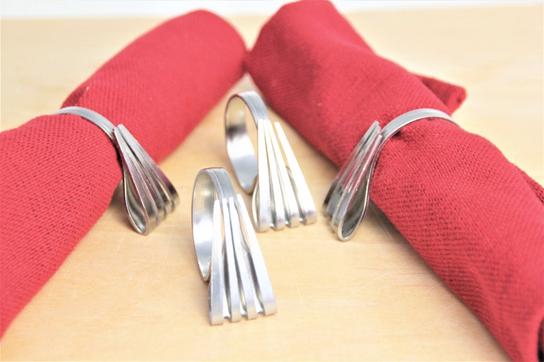 May include: Four silver napkin rings made of stacked metal strips. The rings are shown on red napkins.