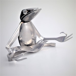 May include: A silver frog sculpture made from spoons and forks, sitting in a meditative pose with its legs crossed and hands resting on its knees.