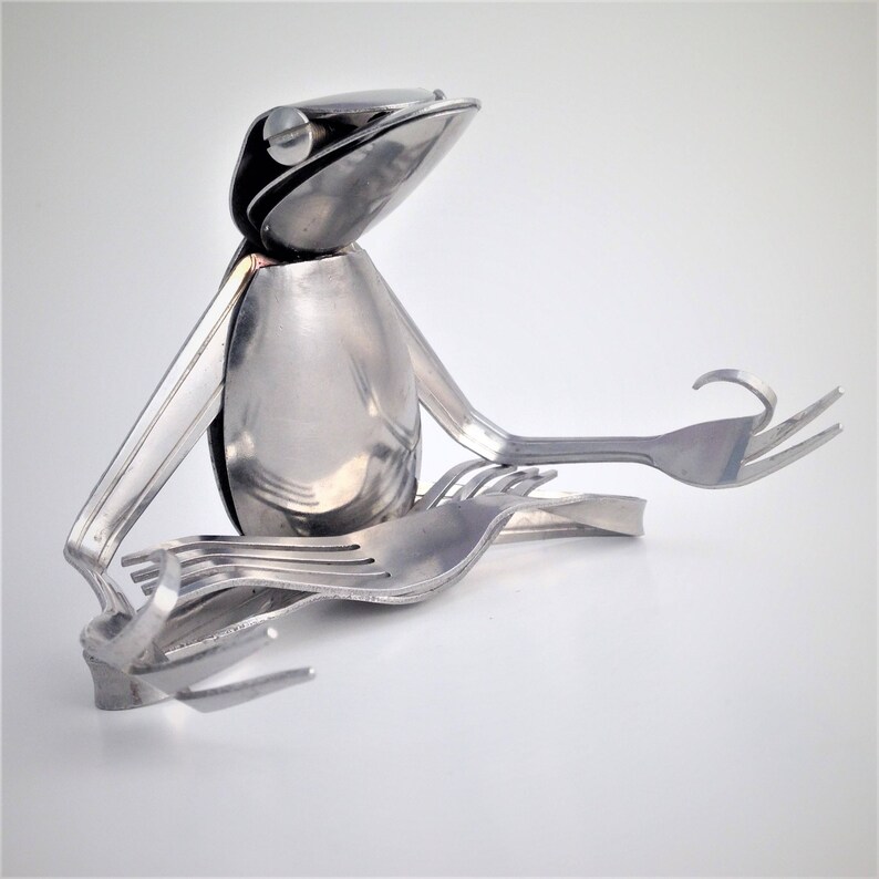 May include: A silver frog sculpture made from spoons and forks, sitting in a meditative pose with its legs crossed and hands resting on its knees.