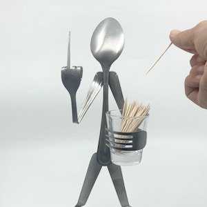 May include: A whimsical sculpture crafted from silverware, featuring a spoon as the head and a fork as an arm. A small glass holds toothpicks, and a hand is holding a toothpick. The sculpture is on a white background.