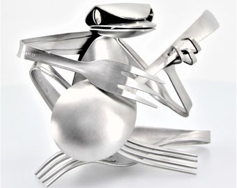 May include: A silver frog sculpture made from recycled silverware, playing a banjo with a fork. The frog's head is formed from a spoon, and the banjo is made from a knife.