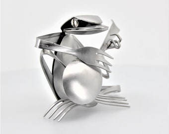 May include: A silver frog sculpture made from recycled silverware. The frog is sitting on its hind legs and holding a guitar made from a fork and spoon.