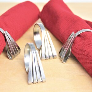 May include: Four silver napkin rings made from repurposed silverware. The rings are designed with a fan-like shape and are shown on a wooden surface with red napkins.
