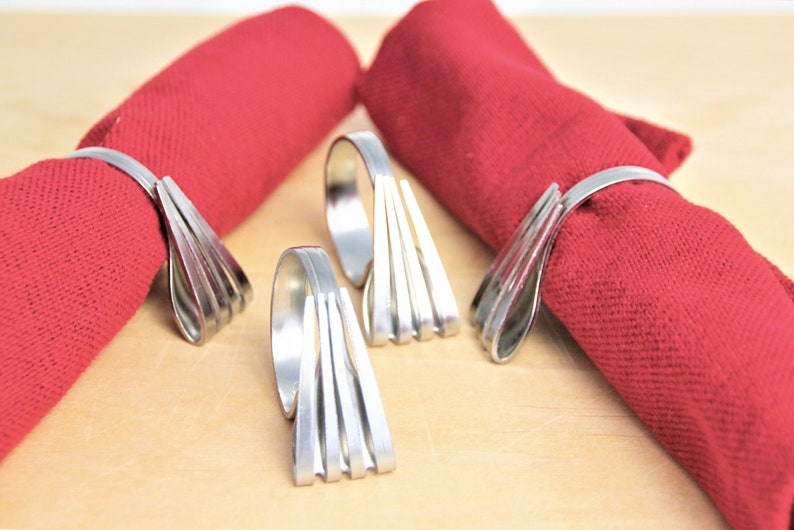 May include: Four silver napkin rings made from repurposed silverware. The rings are designed with a fan-like shape and are shown on a wooden surface with red napkins.
