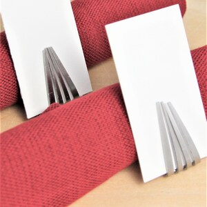 May include: Two white card holders with silver metal holders are placed on top of red napkins. The napkins are rolled up and placed on a wooden surface.