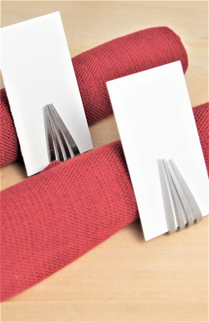 May include: Two white card holders with silver metal holders are placed on top of red napkins. The napkins are rolled up and placed on a wooden surface.