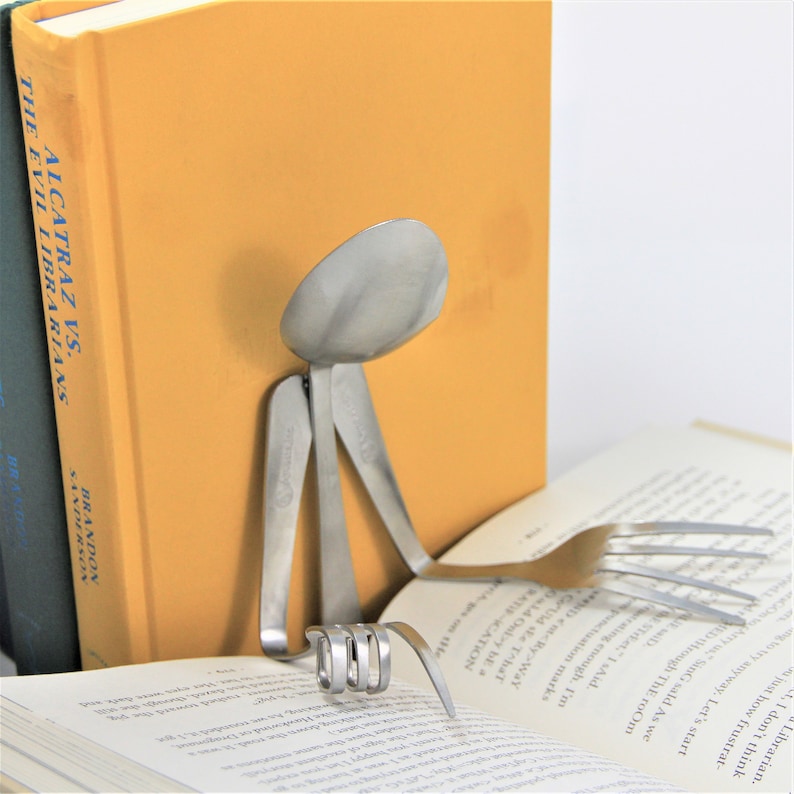 Puede incluir: Una escultura de metal plateado de una persona leyendo un libro. La escultura est&aacute; hecha de cubiertos doblados, incluyendo una cuchara para la cabeza y un tenedor para un brazo. La escultura est&aacute; sentada sobre un libro con el t&iacute;tulo "Alcatraz vs. The Evil Librarians" de Brandon Sanderson.
