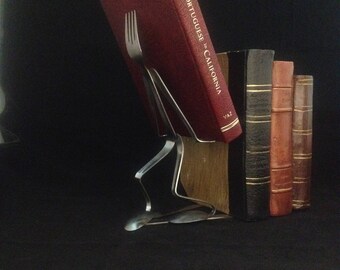 May include: A silver fork and spoon are bent into a shape that holds up a stack of books. The top book is red and has the title "The Portuguese in California" on the spine. The other books are brown and black.
