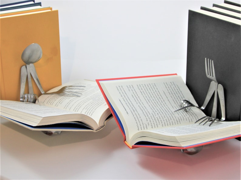 Puede incluir: Dos sujetalibros hechos de cubiertos, con forma de personas, sosteniendo libros abiertos. Un sujetalibros est&aacute; hecho de un tenedor y el otro de una cuchara. Los sujetalibros sostienen libros abiertos con cubiertas rojas y azules.