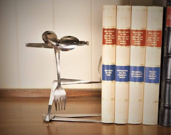 May include: A silver bookend made from bent silverware, including a fork and spoons. The bookend is holding up a set of books with gold lettering on the spines. The books are titled "The History of the Decline and Fall of the Roman Empire" and "The Turn of the Tide".