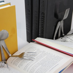 Puede incluir: Un libro con una encuadernaci&oacute;n roja est&aacute; apoyado abierto por una cuchara y un tenedor de plata, formando una figura humana. El libro est&aacute; apoyado sobre dos libros negros con la palabra "RANDOM" impresa en los lomos.