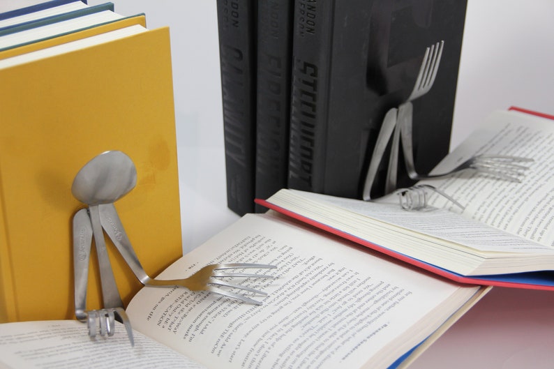 Puede incluir: Un libro con una encuadernaci&oacute;n roja est&aacute; apoyado abierto por una cuchara y un tenedor de plata, formando una figura humana. El libro est&aacute; apoyado sobre dos libros negros con la palabra "RANDOM" impresa en los lomos.