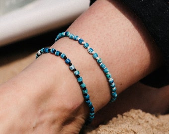 May include: Two beaded anklets in shades of blue and black are worn on an ankle resting on sand. The anklets are made of small, round beads. A white surfboard fin is in the background, and the person is wearing black pants.