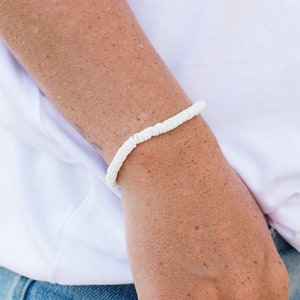 May include: A white shell bracelet on a person's wrist. The bracelet is made of small, white shells that are strung together on a clear elastic cord.