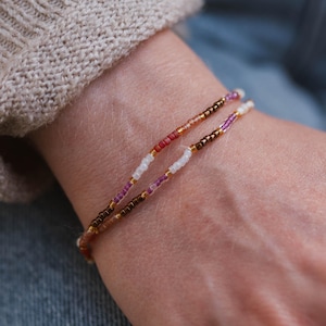 May include: Two delicate beaded bracelets, featuring small, colourful beads in gold, red, white, purple, and brown. The bracelets are worn on a wrist, showcasing the intricate beadwork and design.