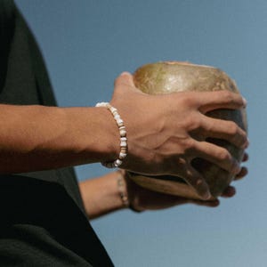 May include: A beaded bracelet with white and tan beads is worn on a wrist. The bracelet is made of small, round beads. The background is a solid light blue. The person is holding a coconut.