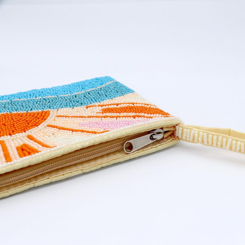 May include: A beaded clutch with a sunset design. The clutch features a cream-colored base with a zipper closure and a wrist strap. The sunset design includes a blue sky, orange sun, and pink and white accents.