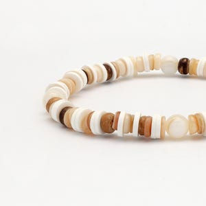 May include: A beaded bracelet featuring a mix of white, tan, and brown circular beads. The bracelet has a natural, earthy aesthetic, with a variety of bead sizes and textures. The beads are strung together to form a flexible band.