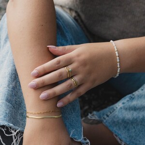 May include: A person wearing a gold chain bracelet, a gold snake chain bracelet, and a white beaded bracelet. The person is also wearing three gold rings on their finger, one with a green stone.