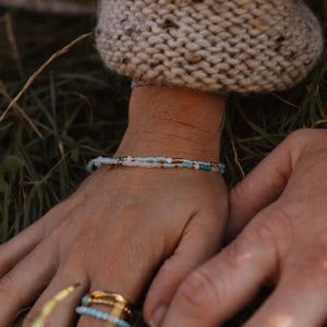 Op de afbeelding: Een delicate kralenarmband met witte, lichtblauwe en gouden kralen. De armband wordt gedragen om een pols met een beige gebreide mouw. Verschillende ringen met turquoise en gouden accenten sieren de vingers.