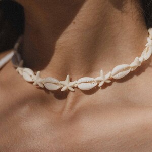 May include: Cream-colored seashell necklace featuring alternating seashells and starfish. The necklace is strung together, creating a delicate and beachy aesthetic. Perfect for adding a touch of coastal charm to any outfit.