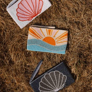 May include: Three beaded clutch bags with seashell and sunset designs. One bag is light gray with a pink seashell. Another features a sunset over blue waves. The third is dark blue with a white seashell.