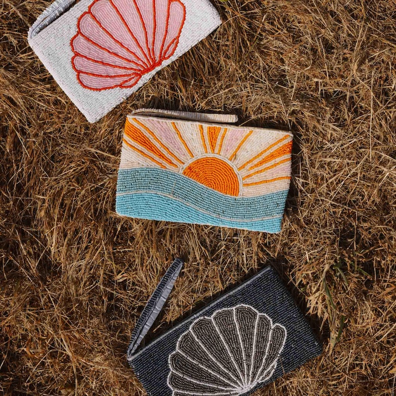 May include: Three beaded clutch bags with seashell and sunset designs. One bag is light gray with a pink seashell. Another features a sunset over blue waves. The third is dark blue with a white seashell.
