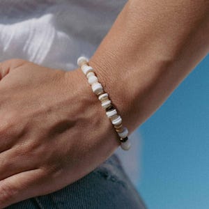 Op de afbeelding: Een armband met witte, bruine en goudkleurige kralen. De armband wordt om een pols gedragen, met een wit overhemd en blauwe achtergrond zichtbaar.