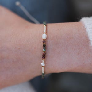 Op de afbeelding: Een kralenarmband met een mix van kleurrijke edelstenen en parels. De armband bevat witte, rode, groene en gouden kralen, geregen met een delicate gouden ketting. De armband is ontworpen om om de pols te worden gedragen.