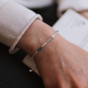 May include: A delicate beaded bracelet featuring a central white stone and a mix of small, colorful beads in shades of teal, blue, and white. The bracelet is strung on a thin, silver-colored cord.
