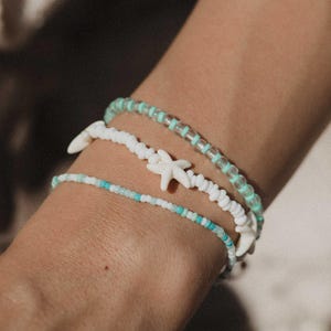 Può includere: Tre braccialetti di perline nei toni del bianco, turchese e trasparente. Un braccialetto presenta ciondoli a forma di stella marina bianchi. I braccialetti sono indossati su un polso, mostrando il design e i colori.