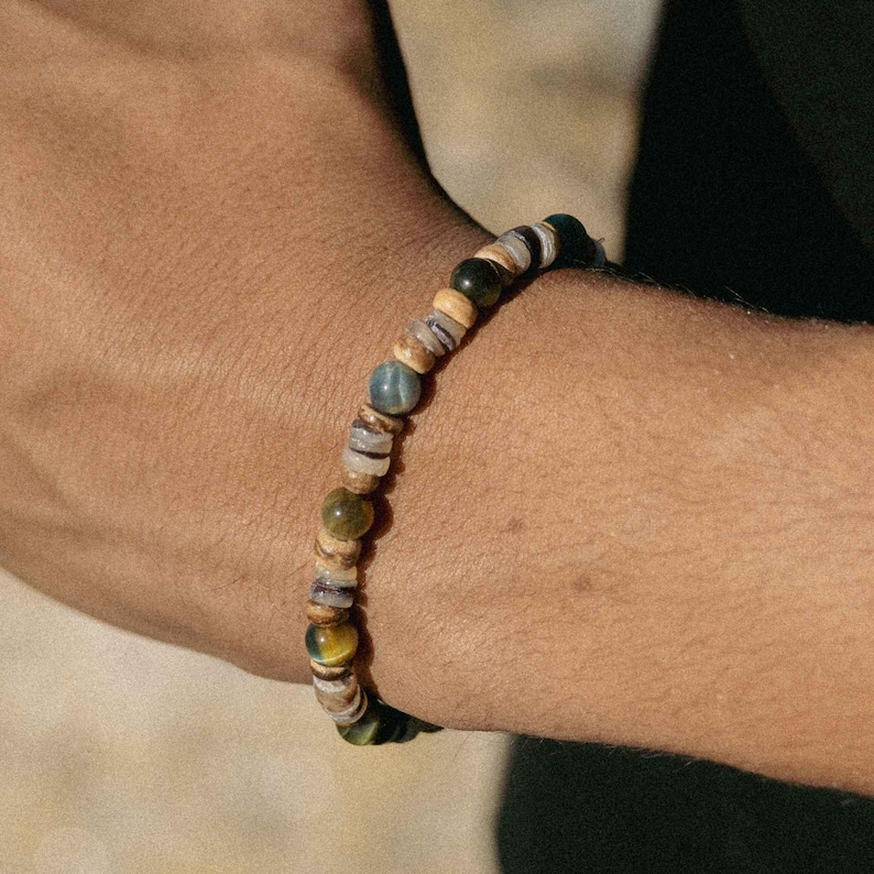 May include: A beaded bracelet featuring a mix of natural stone beads in shades of green, blue, brown, and tan. The bracelet is strung on an elastic cord and is worn on the wrist. The beads vary in size and shape, creating a unique and organic look.