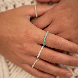 Könnte beinhalten: Ein Satz von vier Perlenringen in verschiedenen Farben. Die Ringe bestehen aus kleinen, runden Perlen und sind auf einem Finger einer Person übereinander gelegt.