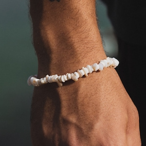 Könnte beinhalten: Ein Armband aus weißen Muschelsplittern. Das Armband besteht aus kleinen, unregelmäßig geformten weißen Muschelteilen, die aneinandergereiht sind. Das Armband wird am Handgelenk getragen.