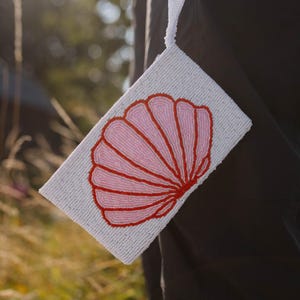 Può includere: Una pochette bianca con perline con un design a conchiglia rosa e rossa. La pochette rettangolare ha un cinturino bianco. Il design a conchiglia è centrato sulla parte anteriore.