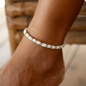 May include: Ankle bracelet featuring small, round, white and pale green beads. The bracelet is worn on the ankle, presenting a simple, beach-inspired design. The beads are strung together, forming a delicate, summery accessory.