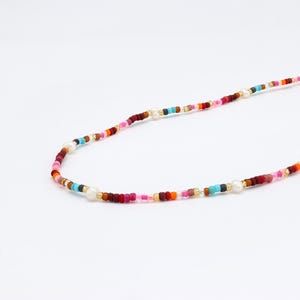 May include: A multi-coloured beaded necklace featuring small, round beads in shades of red, pink, orange, blue, and gold. The necklace also has several white pearl beads. The beads are strung on a thin, flexible cord.