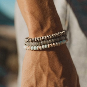 Puede incluir: Tres pulseras de cuentas apiladas en una muñeca. La pulsera superior tiene cuentas moteadas en blanco y negro. La pulsera del medio tiene cuentas grises. La pulsera inferior tiene una mezcla de cuentas azul claro, blancas y marrones.