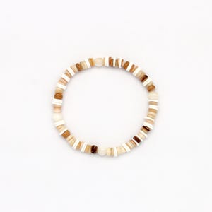 May include: A beaded bracelet featuring a mix of natural-toned beads in shades of white, tan, and brown. The bracelet is circular, with the beads strung together to form a continuous loop. The beads are of varying shapes and sizes.