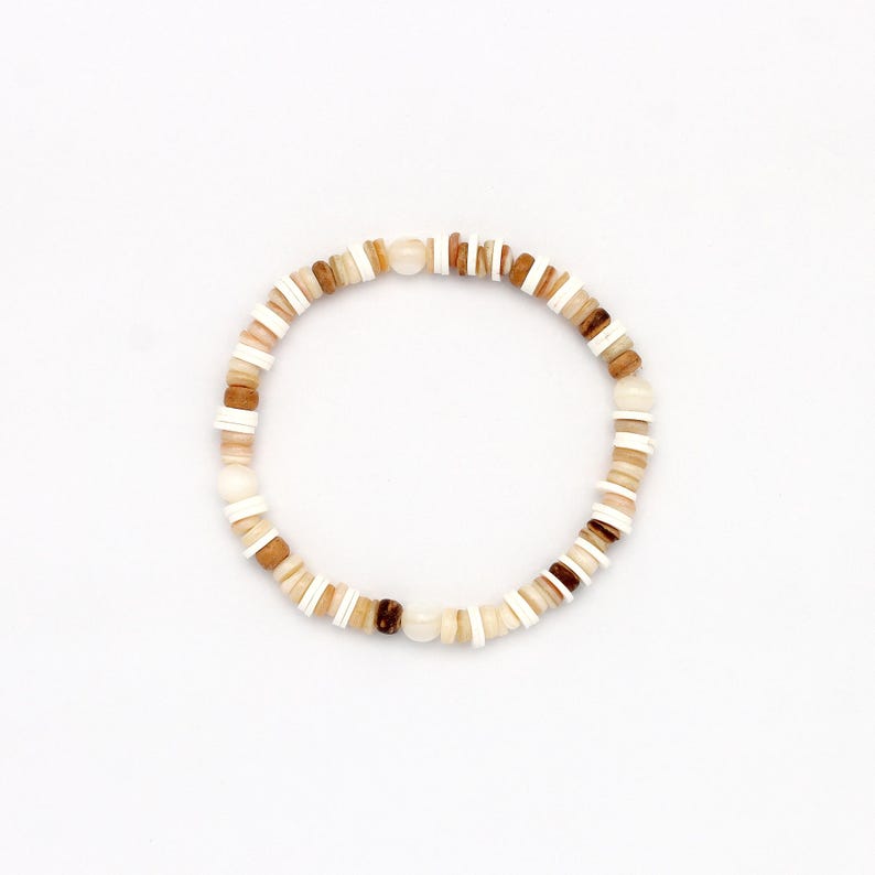 May include: A beaded bracelet featuring a mix of natural-toned beads in shades of white, tan, and brown. The bracelet is circular, with the beads strung together to form a continuous loop. The beads are of varying shapes and sizes.